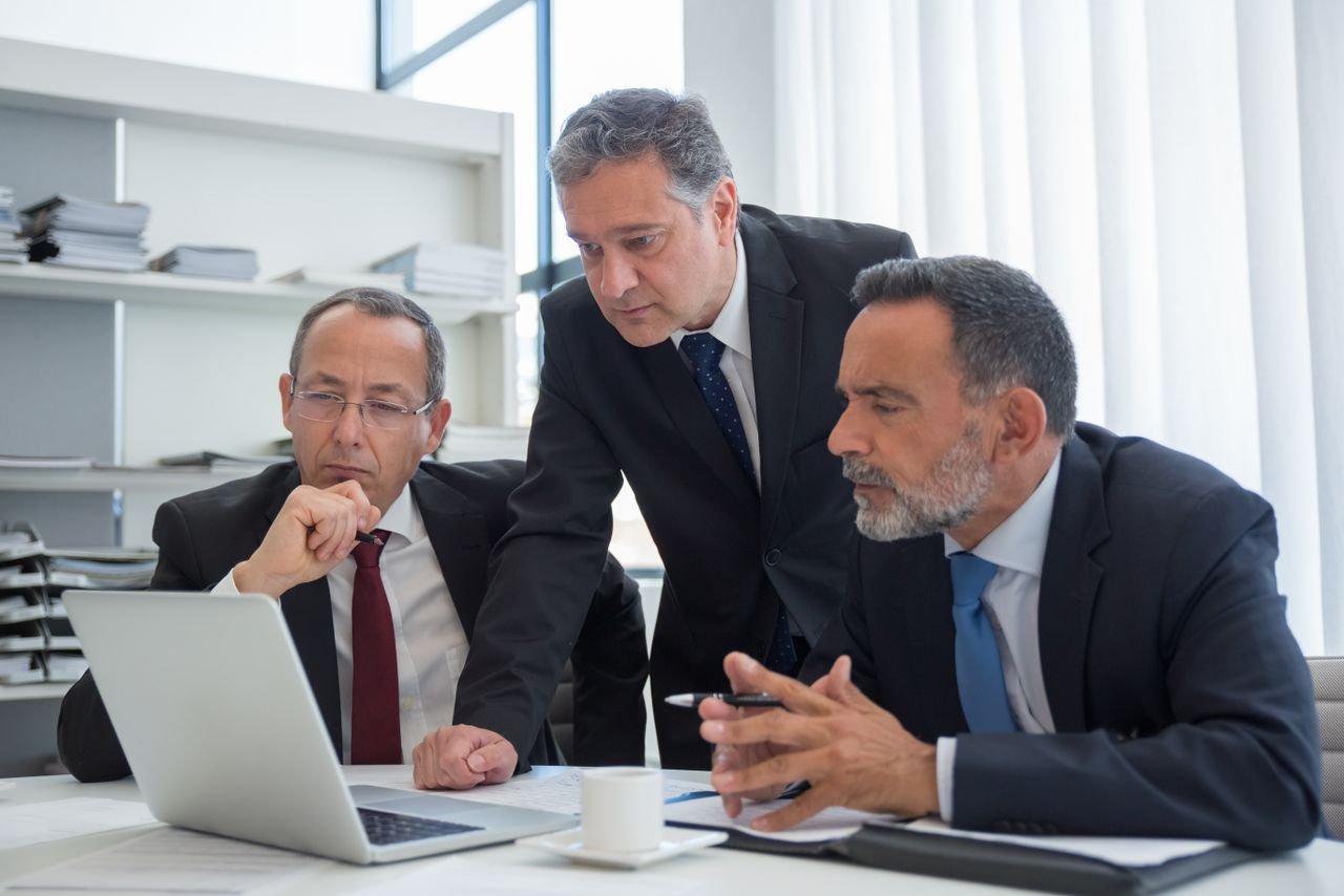 about-03 Three businessmen in suits collaborating in a modern office setting, focused on a laptop.